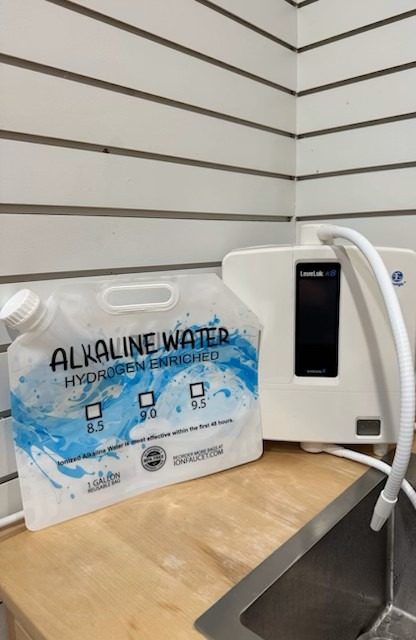 A white countertop water ionizer machine with a flexible hose sits next to a labeled alkaline water bag, featuring pH levels of 8.5, 9.0, and 9.5, on a wooden counter—perfect for enjoying fresh molecular hydrogen water by the stainless steel sink. - Wing Wellness Medical Spa A white countertop water ionizer machine with a flexible hose sits next to a labeled alkaline water bag, featuring pH levels of 8.5, 9.0, and 9.5, on a wooden counter—perfect for enjoying fresh molecular hydrogen water by the stainless steel sink.