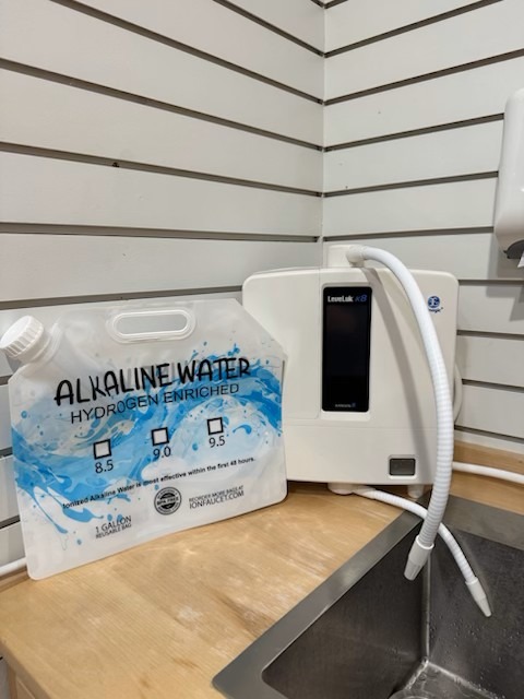 A white countertop water ionizer machine with a flexible hose sits next to a labeled alkaline water bag, featuring pH levels of 8.5, 9.0, and 9.5, on a wooden counter—perfect for enjoying fresh molecular hydrogen water by the stainless steel sink. - Wing Wellness Medical Spa A white countertop water ionizer machine with a flexible hose sits next to a labeled alkaline water bag, featuring pH levels of 8.5, 9.0, and 9.5, on a wooden counter—perfect for enjoying fresh molecular hydrogen water by the stainless steel sink.
