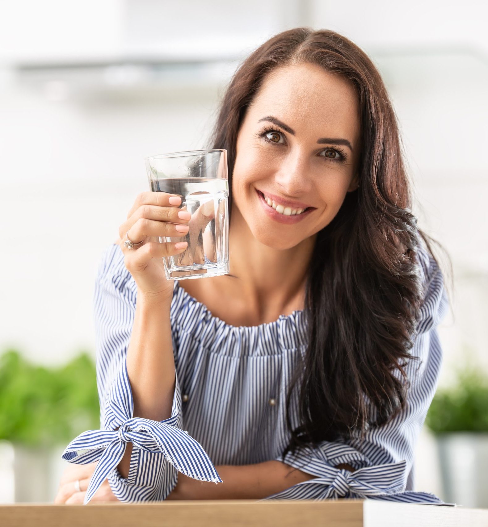 A woman with long dark hair, wearing a blue and white striped blouse, is smiling and holding a glass of water in a brightly lit kitchen with green plants in the background.