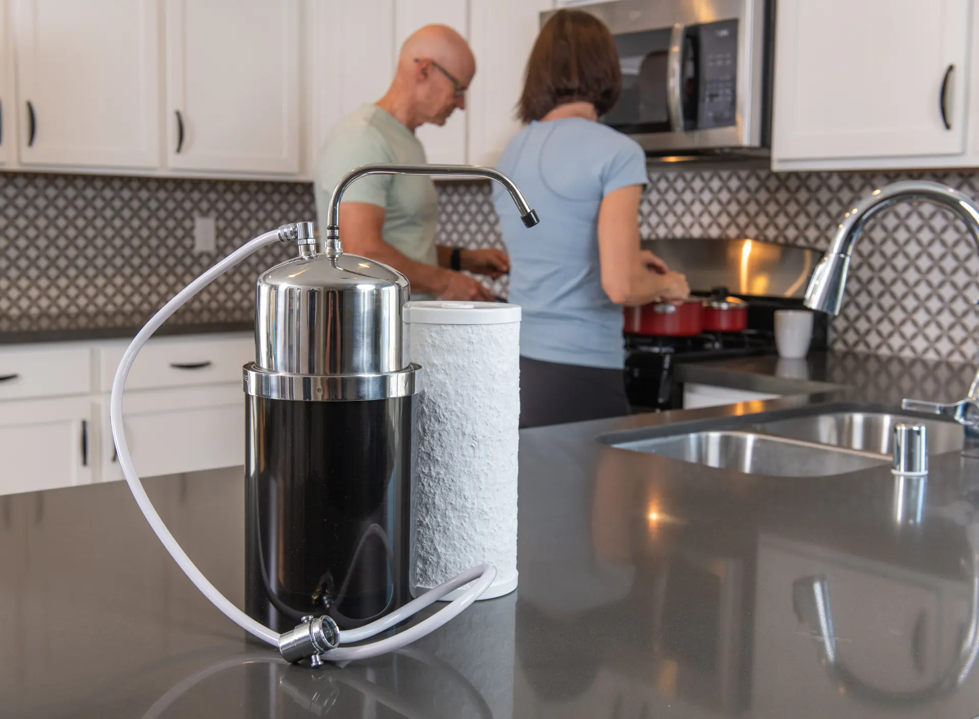 A countertop water filter and a paper towel roll sit on a kitchen island. In the background, a man and a woman cook together by the stove in a modern kitchen.