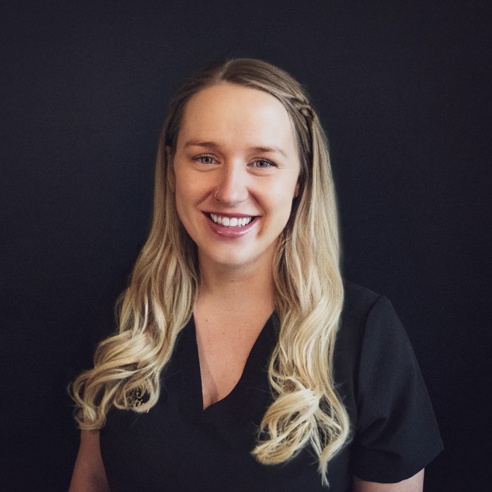 A woman with long, wavy blonde hair wearing a black top is smiling at the camera against a plain, dark background.